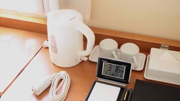 A table with a white electric kettle, two white cups, a digital clock, a white power cord, a black notepad, and a pen.