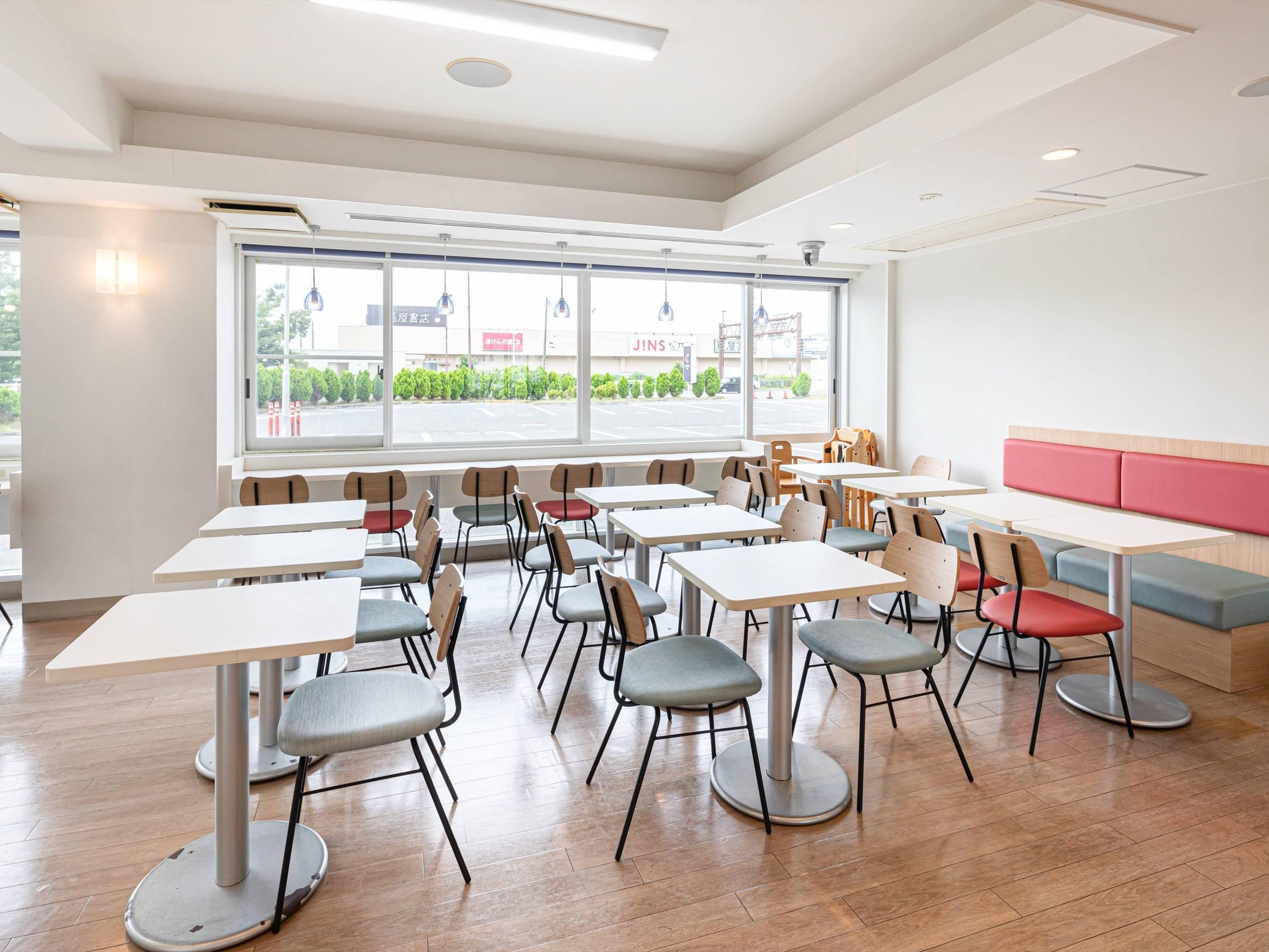 A modern dining area with multiple tables and chairs, large windows with a view of the street, and a red cushioned bench.