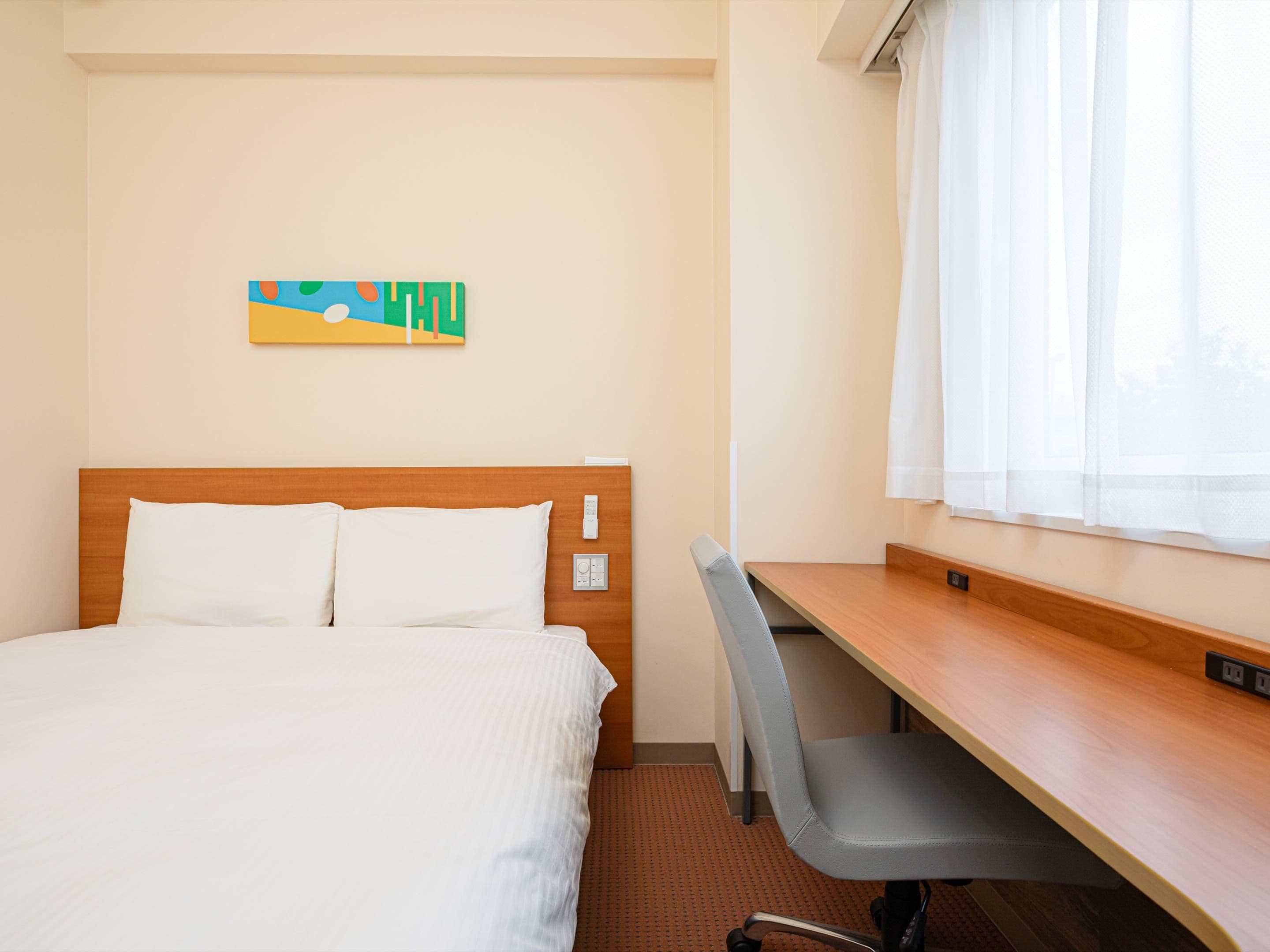 A hotel room with a bed, a desk, and a window with white curtains.