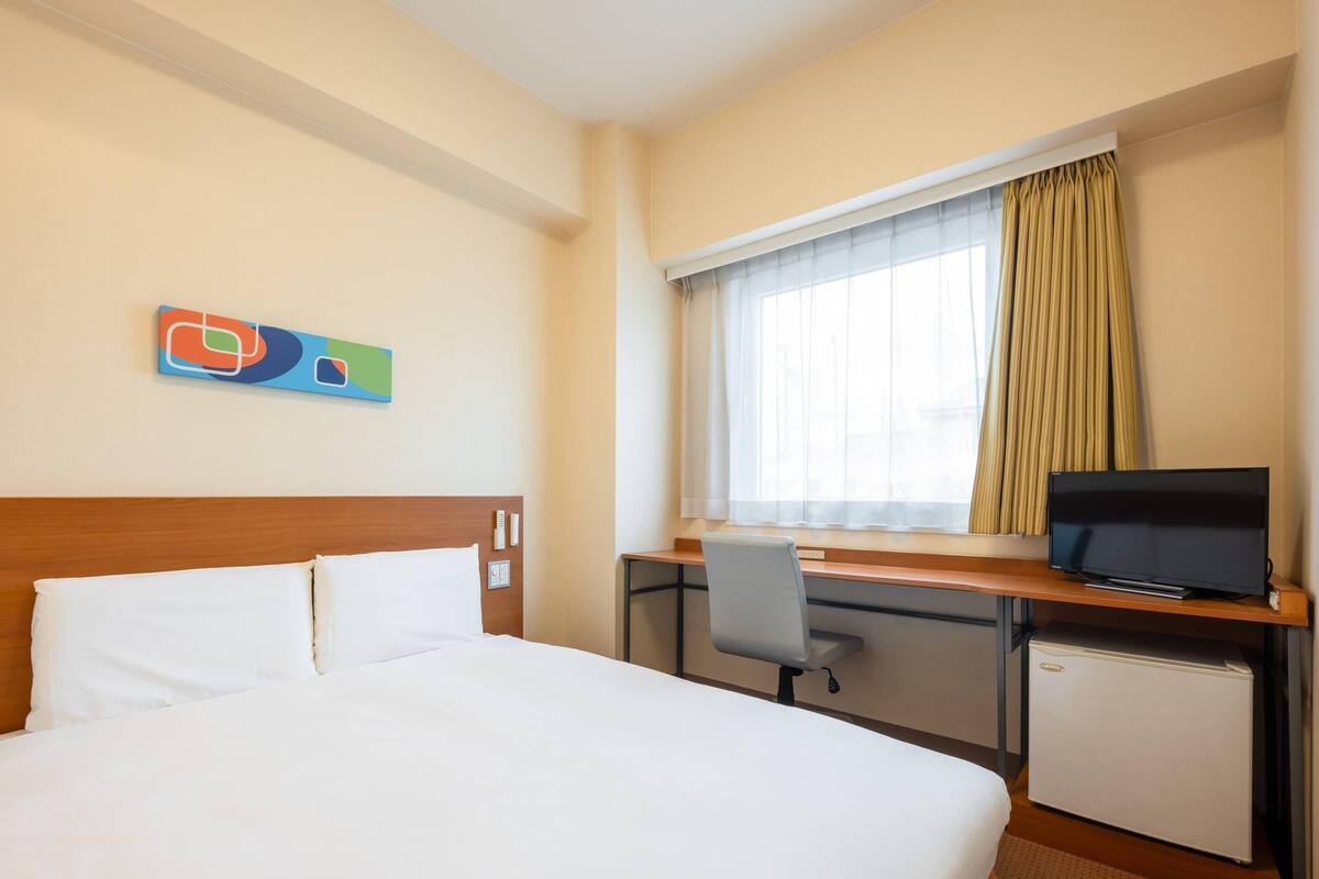 A hotel room with a bed, a desk with a television, a chair, and a window with curtains.