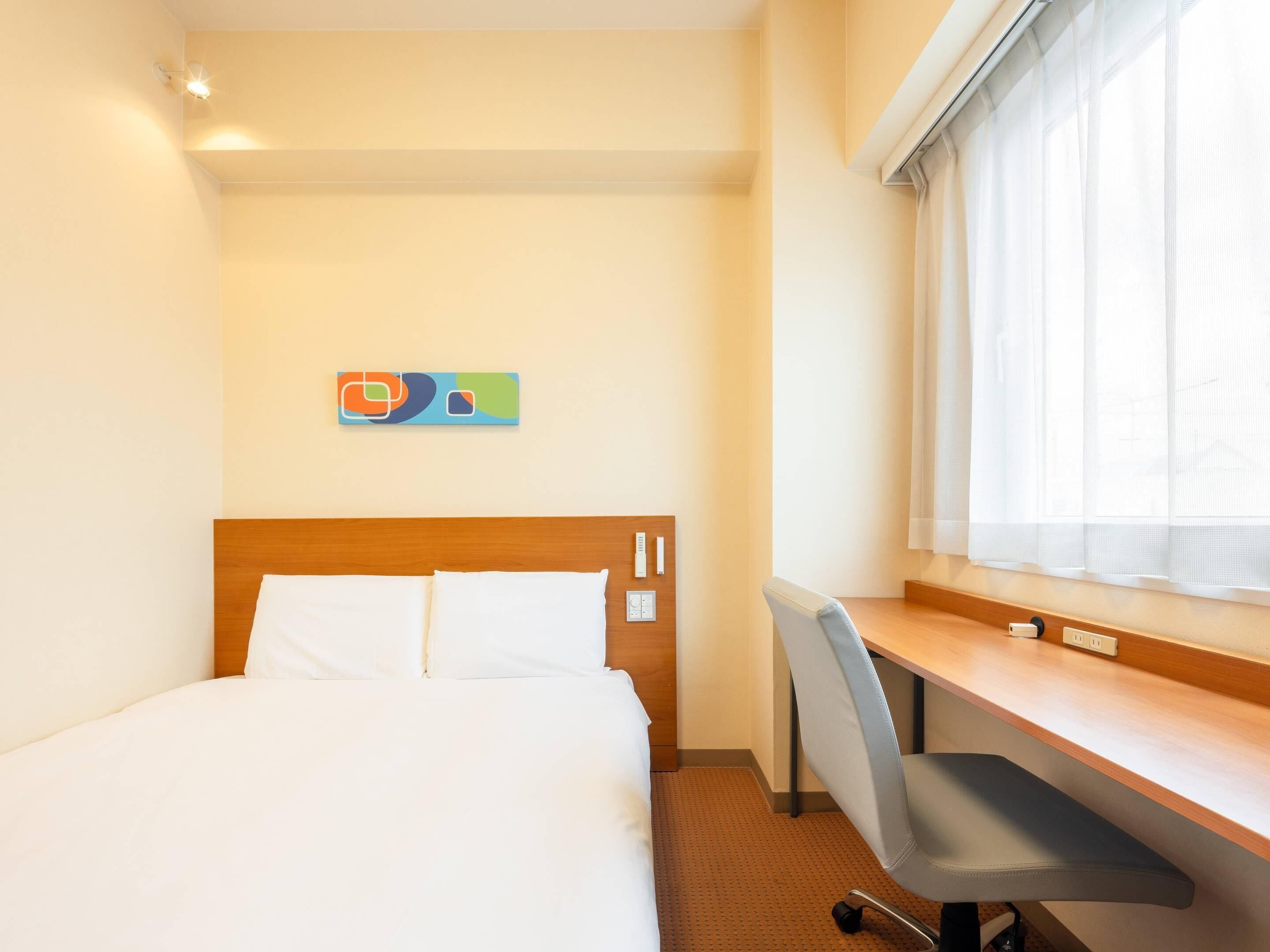 A hotel room with a bed, a desk, and a window with sheer curtains.
