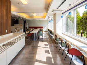 A modern dining area with a long table and chairs, a kitchen area with a refrigerator, and large windows offering a view of trees. - Comfort Inn Zentsuji Inter (Marugame)