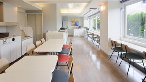 Ein moderner, gut beleuchteter Essbereich mit langen Tischen und Stühlen, eine Küche im Hintergrund und große Fenster mit Blick auf Grünfläche.