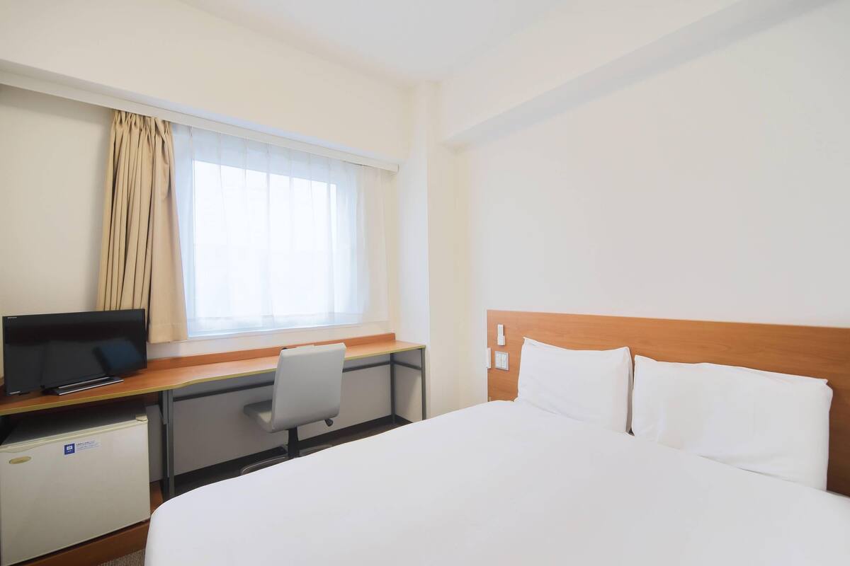 A hotel room with a bed, a desk with a computer, a chair, a small refrigerator, and a window with curtains.