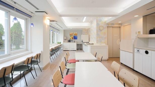 Uma área de refeições moderna com mesas e cadeiras brancas, uma cozinha ao fundo e amplas janelas que proporcionam luz natural.
