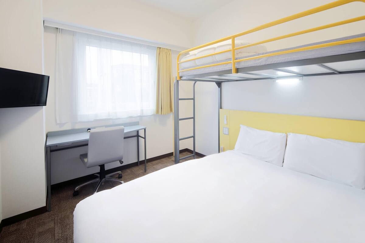 A hotel room with a bunk bed, a desk with a chair, a television, and a window with curtains.
