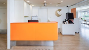 Ein Empfangsbereich mit einem leuchtend orangefarbenen Empfangstresen, einer Kaffeemaschine und einem großen Fenster, das natürliches Licht hereinlässt.