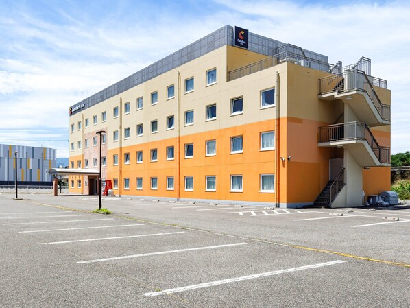 Un hotel de varios pisos con fachada plana, múltiples ventanas y un estacionamiento al frente.