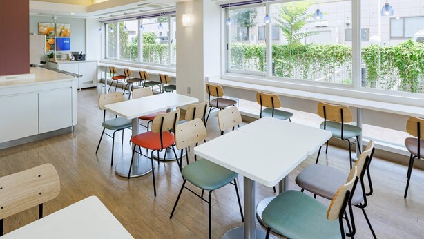 A modern dining area with white tables and chairs, large windows with a view of greenery, and a kitchen area in the background. - Comfort Inn Kofu Isawa (Fuefuki)