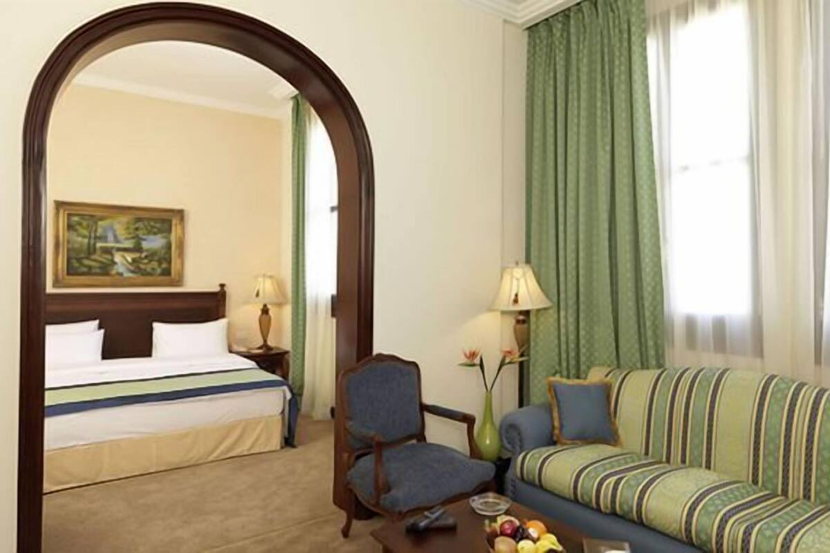 Habitación de hotel con cama, sofá, sillón, una mesa con frutas, una lámpara y un cuadro en la pared.