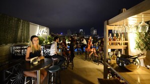 Zona de bar al aire libre con mesas y sillas, una barra con botellas y una vista del horizonte de la ciudad al fondo.