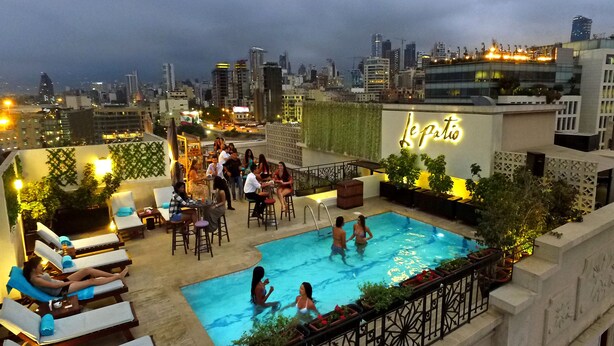 Área de piscina en la azotea con sillones de descanso y un bar, rodeada de vistas a la ciudad y vegetación.
