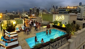 A rooftop pool area with lounge chairs and a bar, surrounded by city views and greenery.