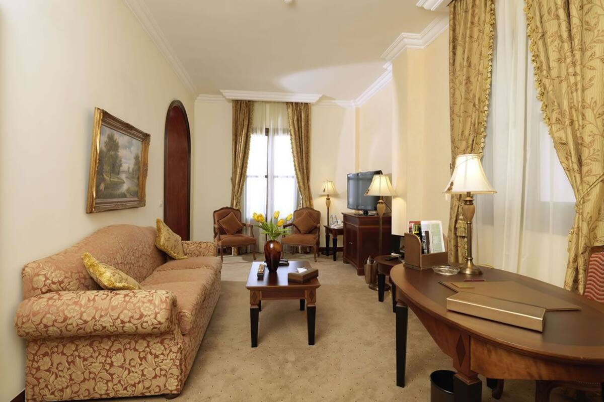 Habitación de hotel con sofá, sillón, mesa de centro, televisor y escritorio con lámpara.