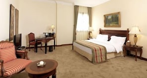 A hotel room with a bed, a desk with a computer, a chair, a small table with a decorative item, and a painting on the wall.