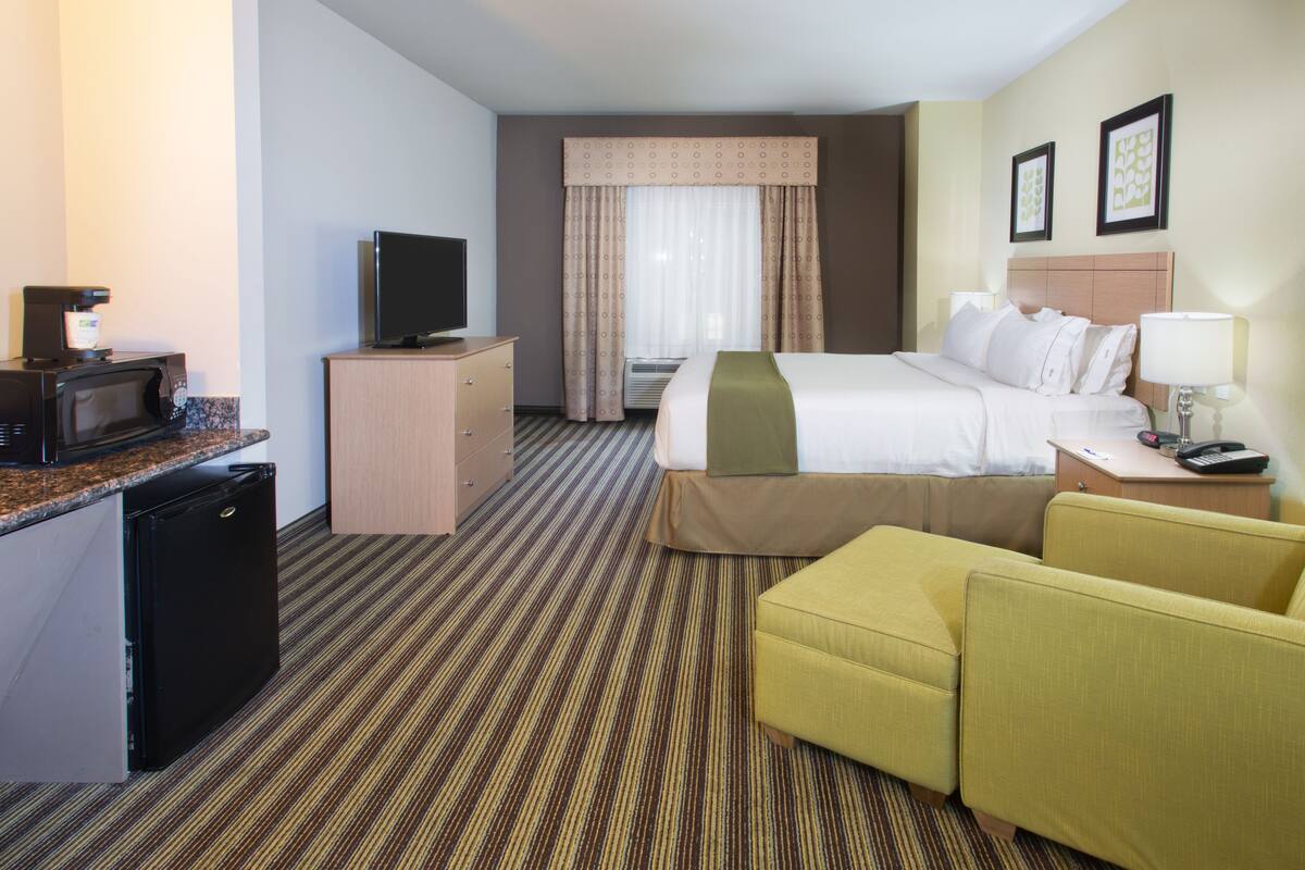A hotel room with a bed, a desk with a computer, a television, a microwave, a small refrigerator, and a seating area with two chairs.