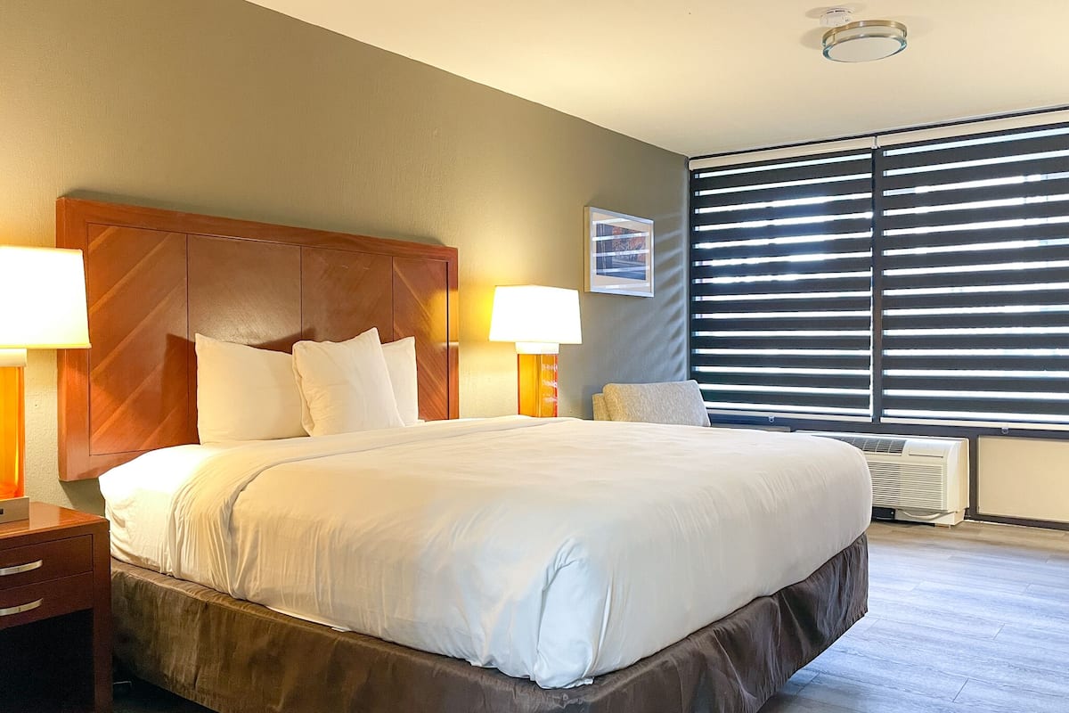 Une chambre d’hôtel avec un grand lit, deux lampes de chevet, une table de chevet, une fenêtre avec des stores et un tableau encadré au mur.