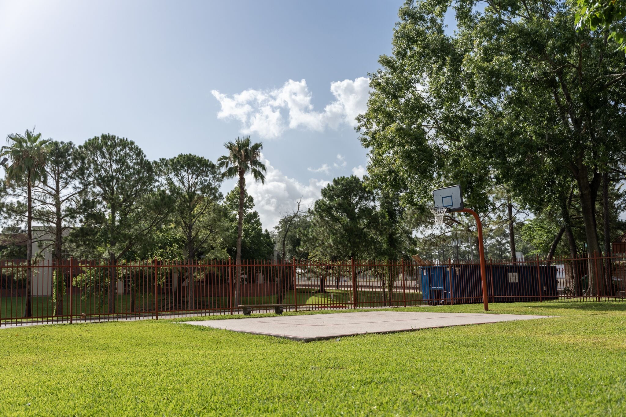 A fenced park with a basketball court, trees, and a clear sky.