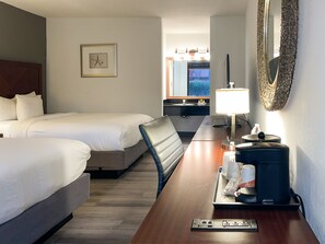 Quarto de hotel com duas camas, uma mesa de trabalho com abajur, um espelho e uma cesta fixada na parede.