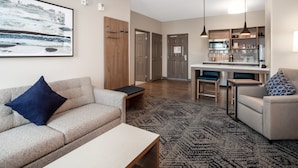 A modern living room with a sofa, coffee table, and a rug. There is a kitchen area with a bar and pendant lights. - Candlewood Suites Grand Junction by IHG (Grand Junction)