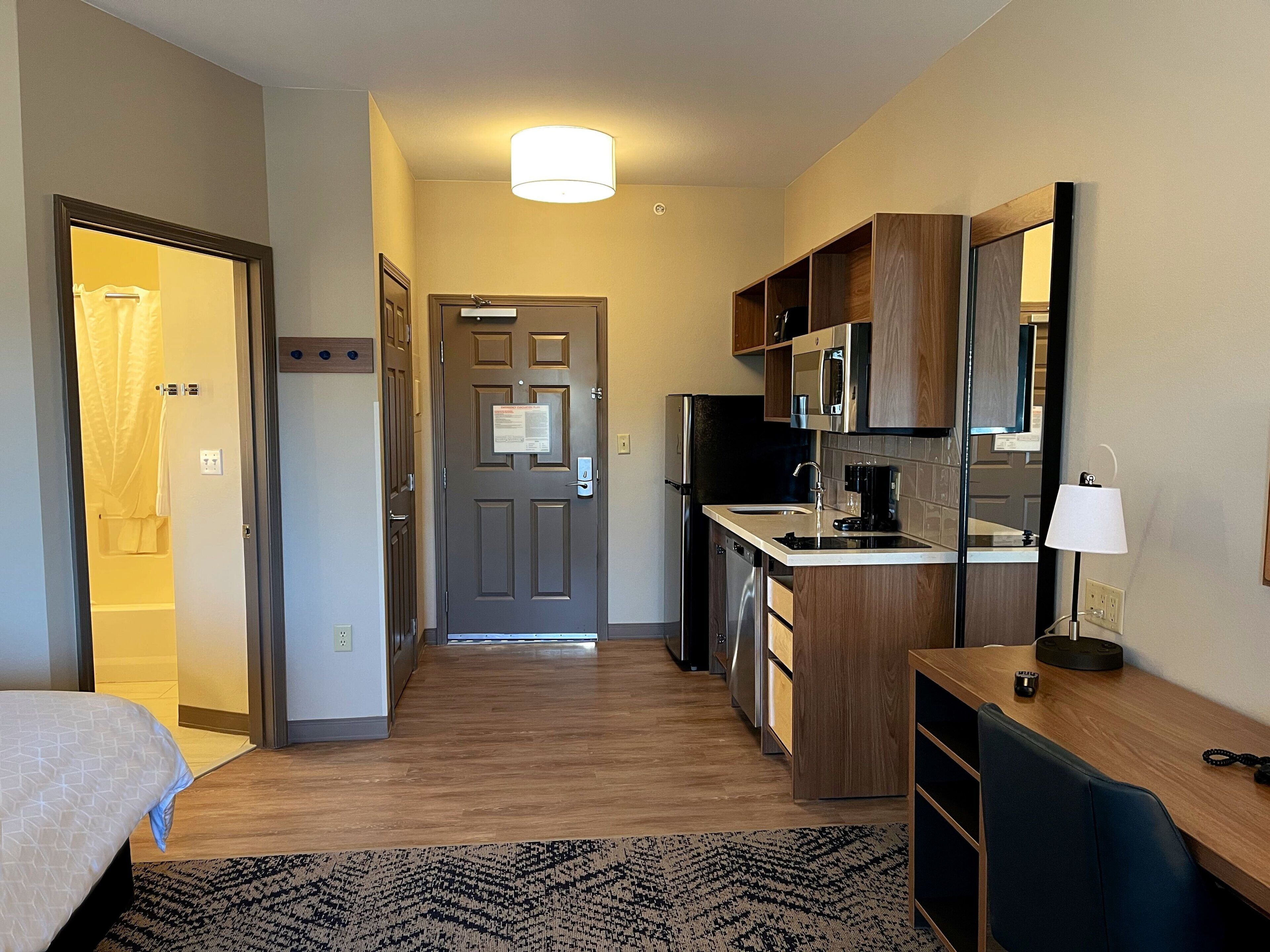 Habitación de hotel compacta con cama, baño con cortina de ducha, kitchenette con microondas y cafetera, y un escritorio pequeño con lámpara.