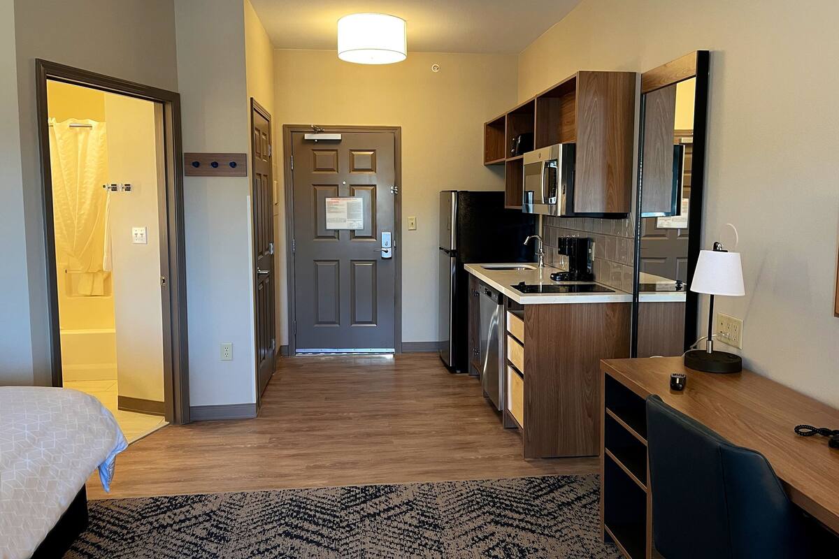 A compact hotel room with a bed, a small kitchenette, a bathroom with a shower curtain, and a hallway leading to the door.