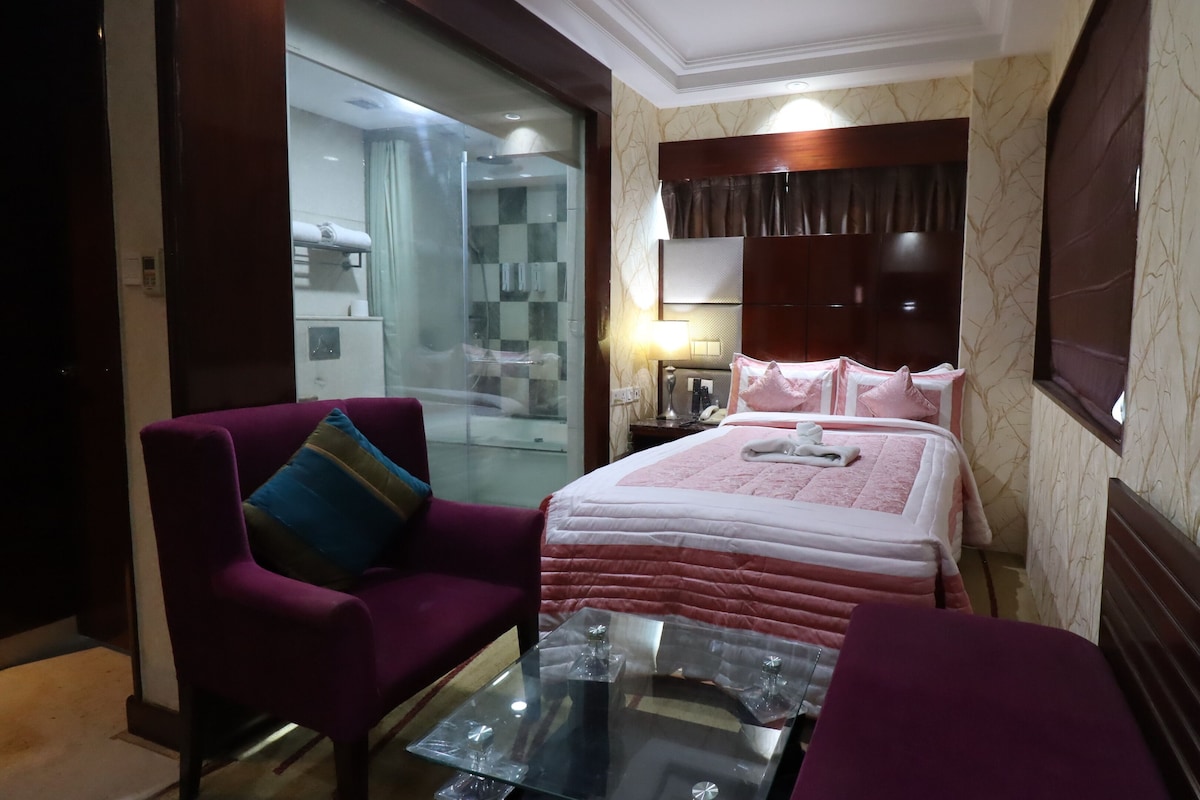 A hotel room with a bed, a glass-enclosed shower, a purple armchair, a glass coffee table, and a nightstand with a lamp.