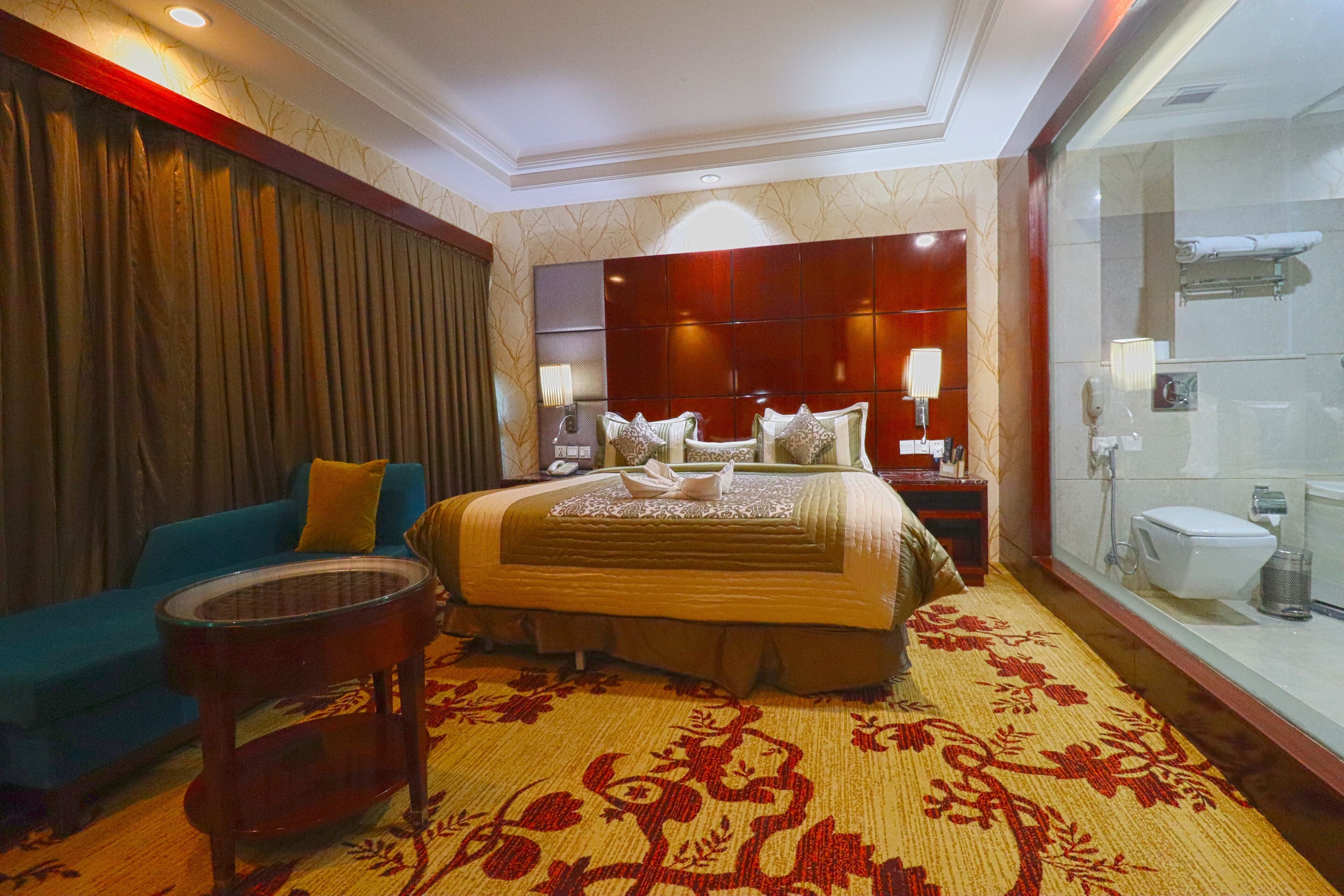 A hotel room with a large bed, a blue sofa, a small round table, and a bathroom with a glass shower enclosure.