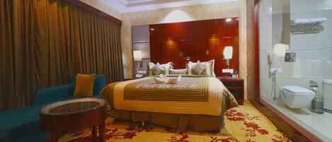 A hotel room with a large bed, a blue sofa, a small round table, and a bathroom with a glass shower enclosure.