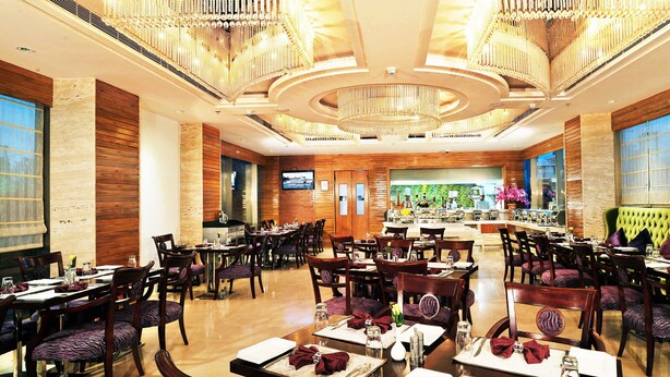 A spacious hotel dining area with multiple tables and chairs, a large chandelier, and a television screen on the wall. - Skycity Hotel (Gurugram)