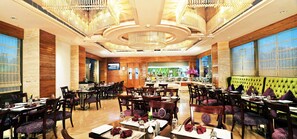 A spacious hotel dining area with multiple tables and chairs, a large chandelier, and a television screen on the wall. - Skycity Hotel (Gurugram)