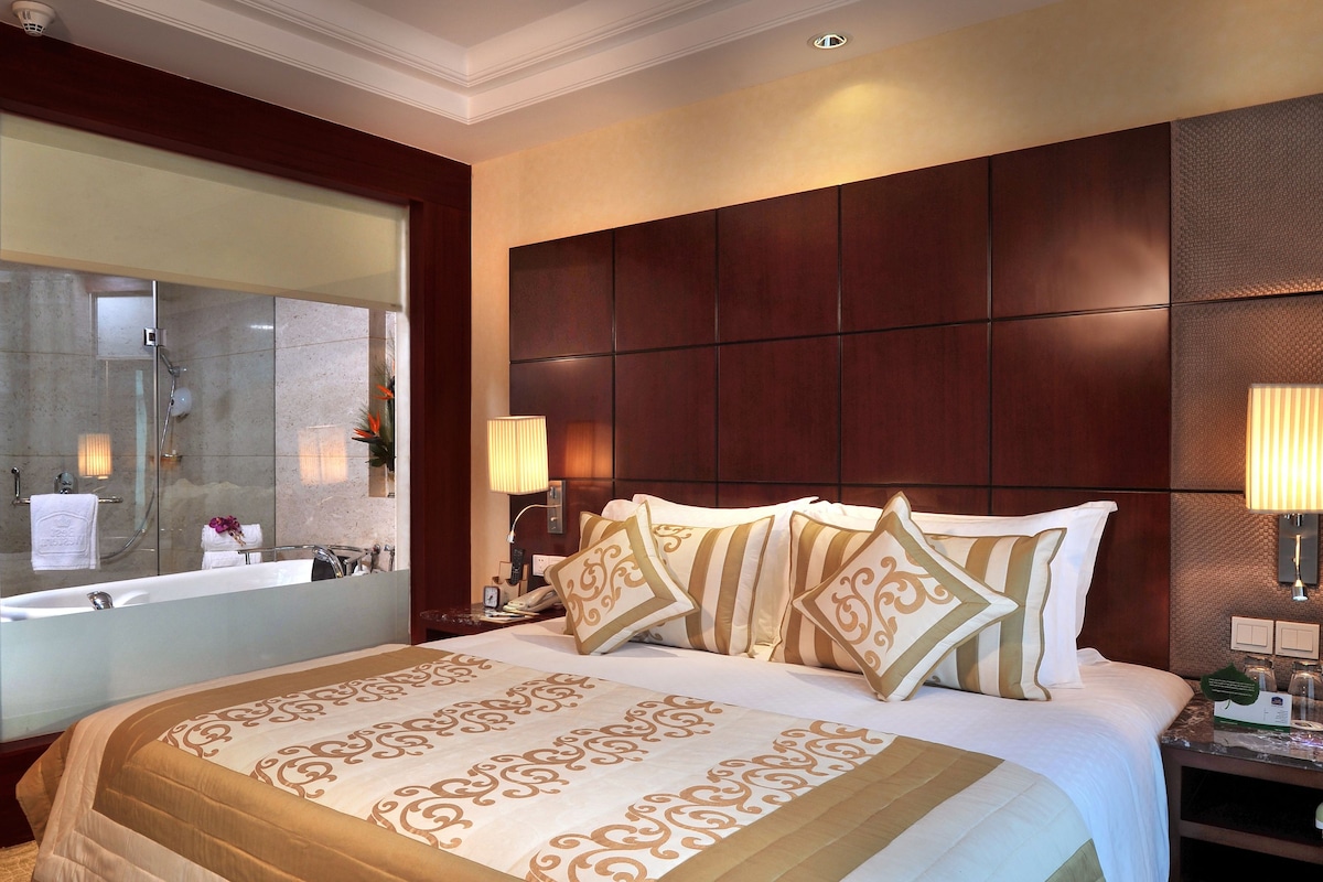 A hotel room with a large bed, a wooden headboard, a bathroom with a bathtub, and a vanity with a mirror.