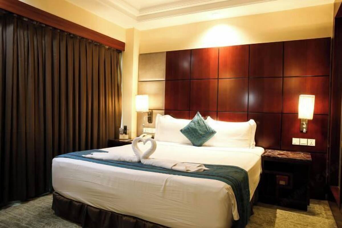 Habitación de hotel con una cama grande, cabecera de madera, dos lámparas de noche y una toalla con forma de corazón sobre la cama.