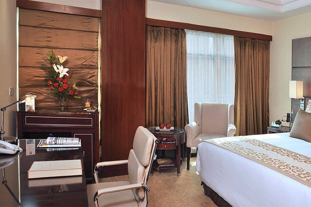 Habitación de hotel con una cama grande, un escritorio con teléfono, una silla, una mesita y una planta decorativa.