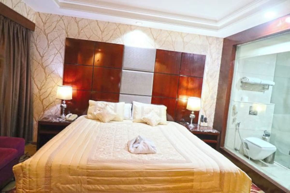 A hotel room with a large bed, two bedside tables, a nightstand, a glass shower enclosure, and a red armchair.