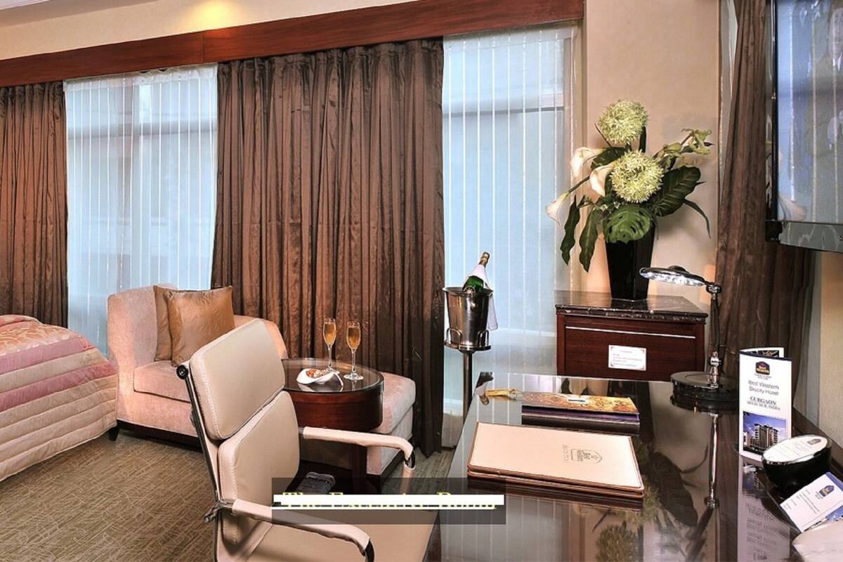 Habitación de hotel con una cama grande, un escritorio con una silla, una mesita con dos copas de champán y un florero con flores.