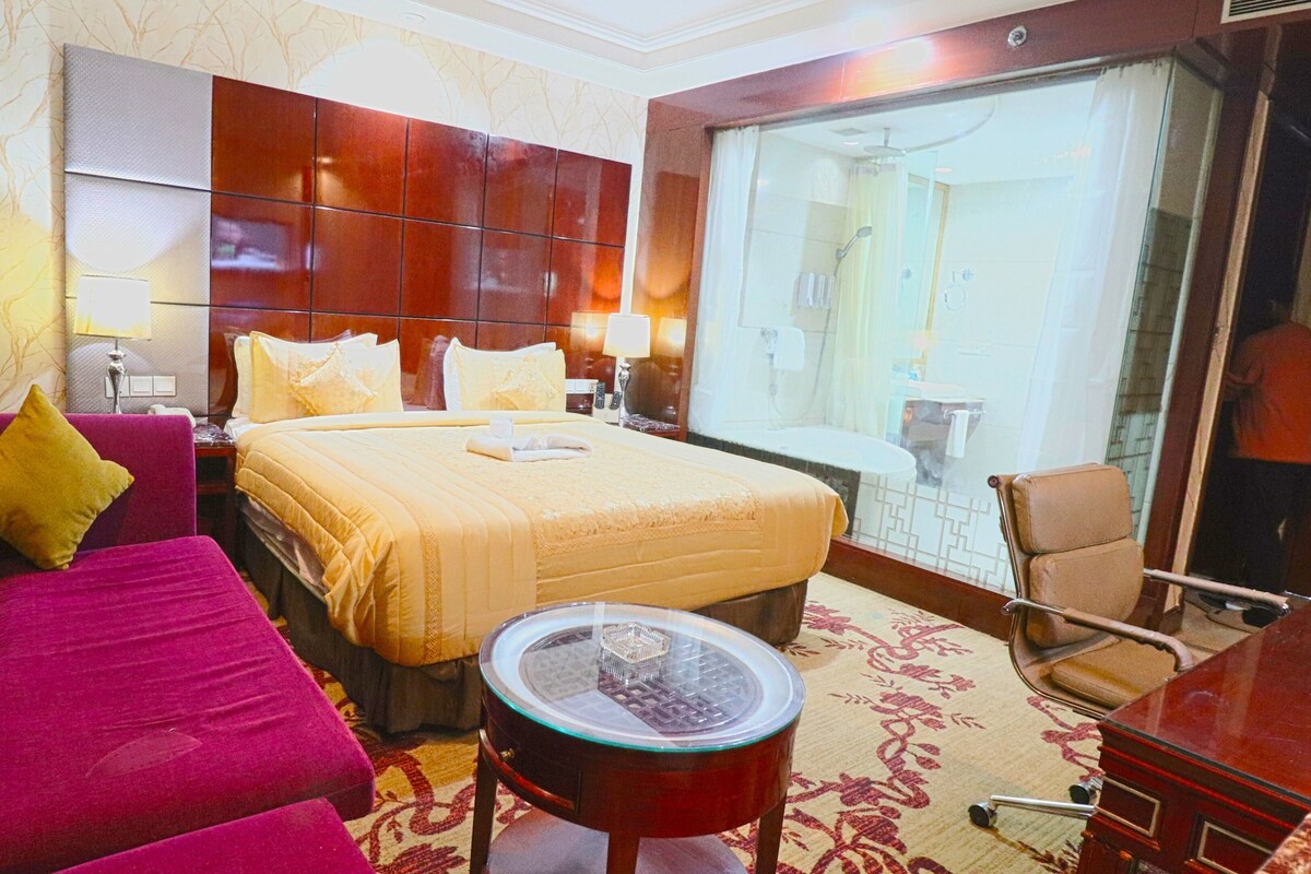 A hotel room with a bed, a sofa, a small table, a chair, and a bathroom visible through a glass door.