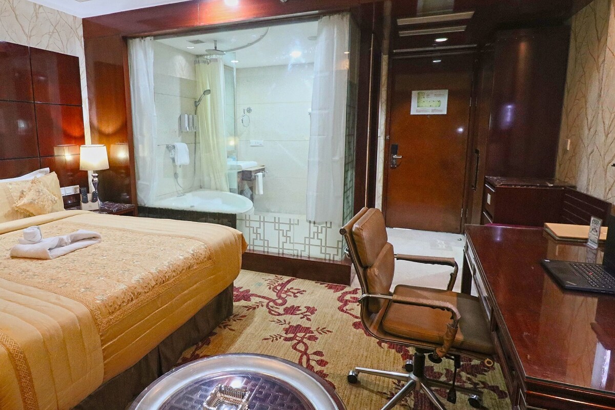 A hotel room with a large bed, a desk with a laptop, a chair, a shower area, and a bathroom.