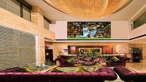 A spacious lobby with a large screen displaying a cricket match, plush seating, a reception desk, and a modern chandelier. - Skycity Hotel (Gurugram)