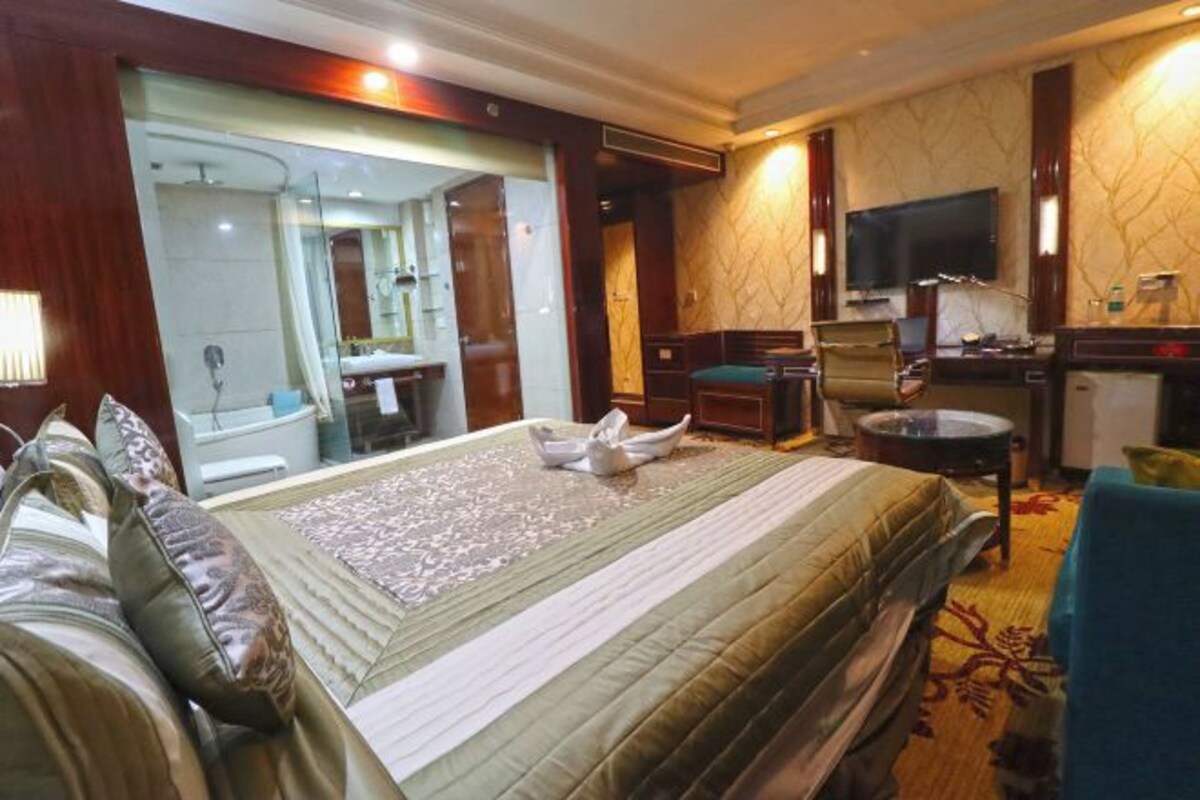 A hotel room with a bed, a desk, a television, and a bathroom visible through a glass door.