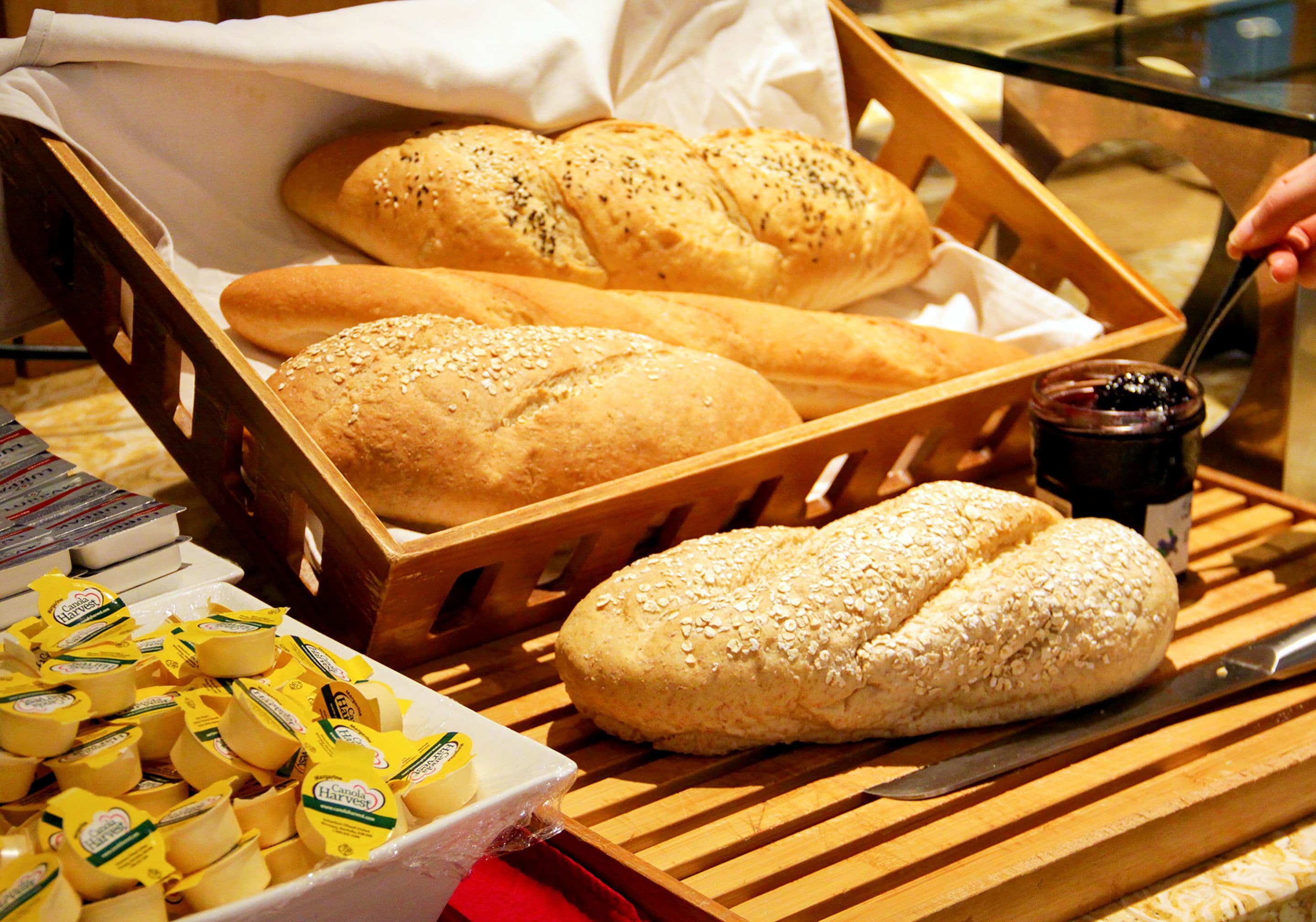 A wooden basket containing various types of bread and a jar of jam on a wooden table.