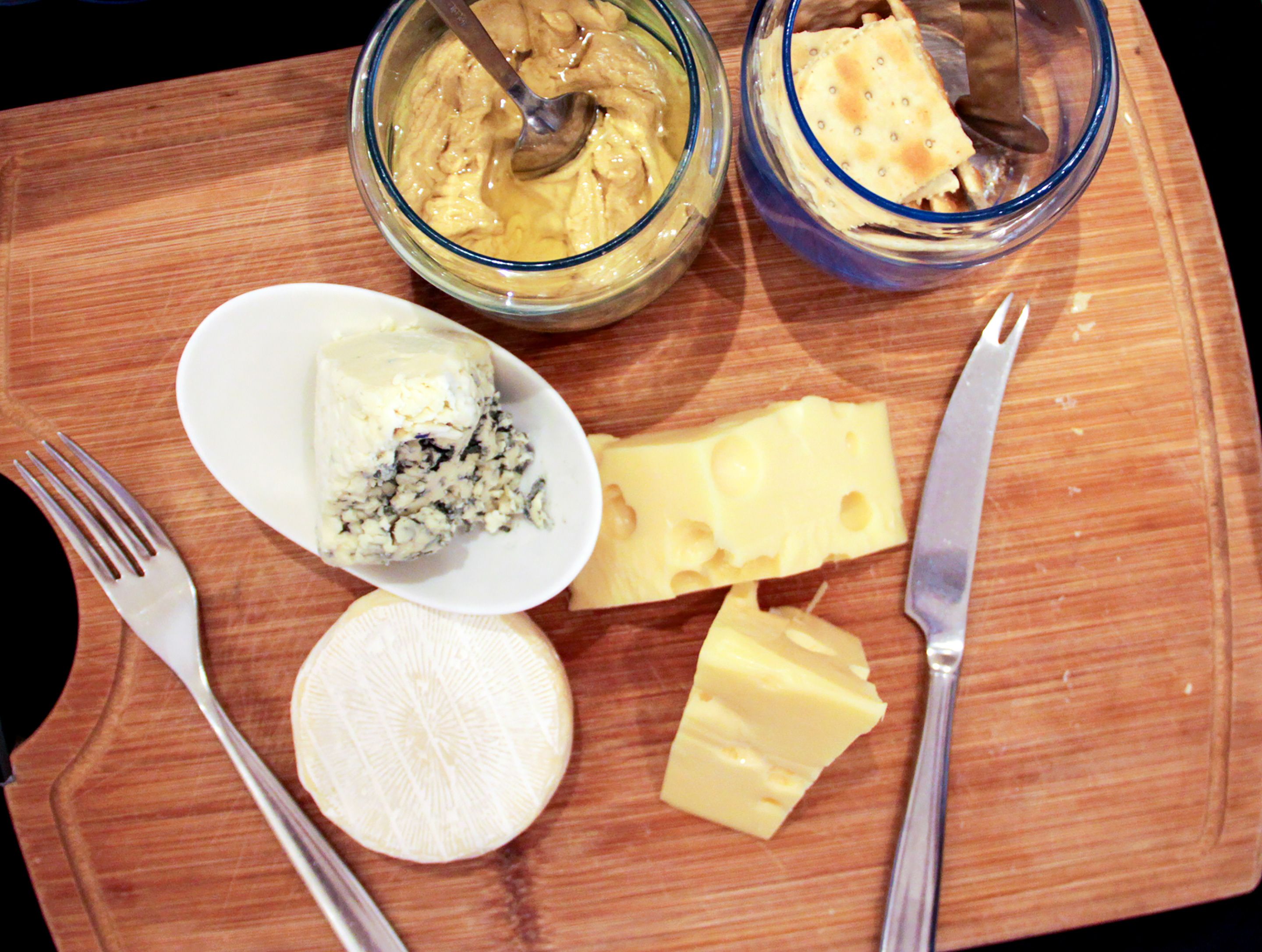 Ein Holzschnittbrett mit einem weißen Teller, der ein Stück Blauschimmelkäse, ein Stück Gelbkäse und ein Stück Weichkäse enthält. Dazu liegen zwei Schalen mit cremiger Substanz und ein Stück Brot bereit.