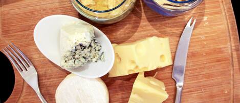 Ein Holzschnittbrett mit einem weißen Teller, der ein Stück Blauschimmelkäse, ein Stück Gelbkäse und ein Stück Weichkäse enthält. Dazu liegen zwei Schalen mit cremiger Substanz und ein Stück Brot bereit.