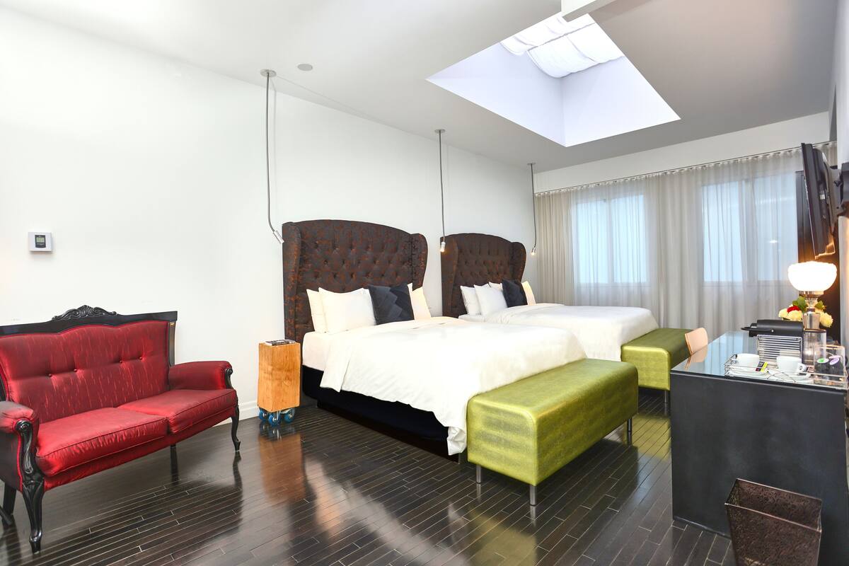 A hotel room with a large bed, a red sofa, a green bench, and a desk with a lamp.