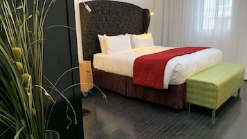 A modern hotel room with a dark wood headboard, a large bed with white and red bedding, a green bench, and a potted plant with tall grass.