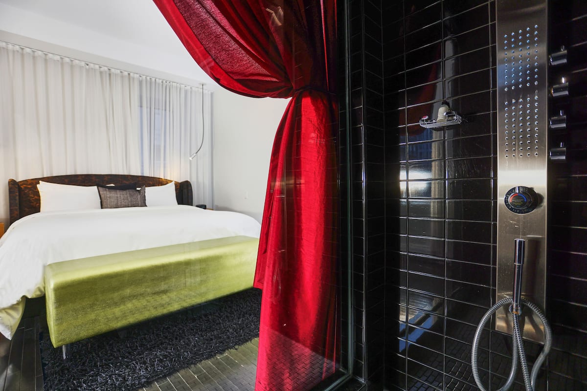 A hotel room with a bed, a green bench, a red curtain, and a shower area with a control panel.