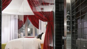 Ein modernes Badezimmer mit Duschkabine aus Glas, einem sichtbaren Bett mit roten Vorhängen und einem Fliesenboden.