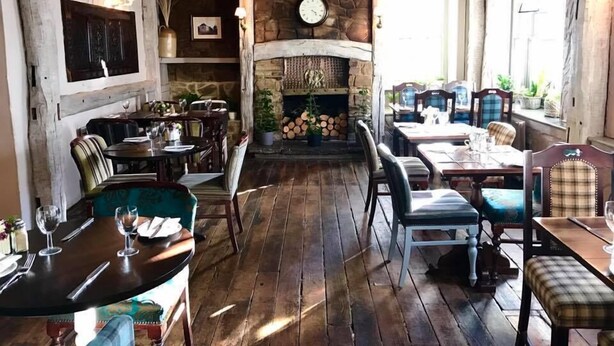 Un interior rústico con pisos de madera, paredes de piedra y una chimenea. Mesas y sillas dispuestas para comer.