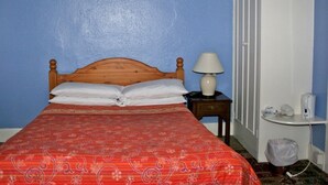 Una cama individual con cabecera de madera, colcha estampada roja, almohada blanca, lámpara de noche, mesita de noche de madera y un bote de basura blanco.