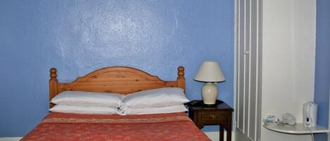 A single bed with a wooden headboard, a red patterned bedspread, a white pillow, a bedside lamp, a small wooden nightstand, and a white trash bin.